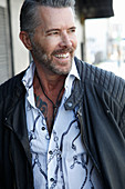 A man with a bread wearing a white, patterned shirt and a leather jacket