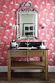 Washstand with marble top below silver-framed mirror on wall with flamingo-patterned wallpaper