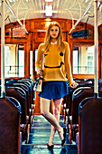 A young blonde woman wearing a smiley jumper and a blue skirt standing on a bus
