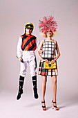 A blonde woman wearing a checked dress and a headpiece with a man in a jockey's outfit jumping behind her