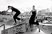 A young woman wearing a black party dress and a young man with a skateboard wearing leisurewear (black-and-white shot)