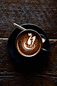 A coffee in a black cup and saucer, with a white milk swirl on a wooden background.
