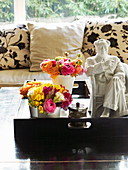 Pillows on sofa, pots with bouquets of flowers and statue on wooden tray in foreground