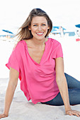 Blonde Frau in pinkfarbener Bluse und Jeans im Sand sitzend