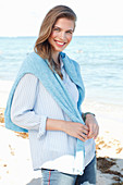 A young woman by the sea wearing a light blue shirt, jumper and jeans