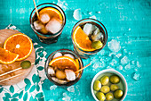 Three glasses of iced vermouth with green olives and orange slices on a turquoise surface
