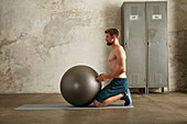 A young man kneeling in front of a gym ball