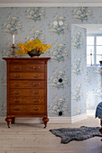 Antique tallboy in bedroom with vintage-style, pale blue, floral wallpaper