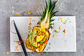A pineapple bowl with oriental noodles, vegetables and avocado in a fresh pineapple on a white marble platter