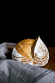 Linen napkin placed on timber tabletop near huge loaf of freshly baked bread against black background