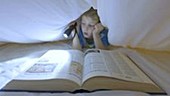 Boy reading book under sheet with torch