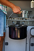 A man holding a pot in front of a gas stove in a camper van