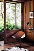 Schalenstuhl aus Holz neben geöffneter Schiebetür zum verwunschenen Gartenbereich