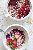 Two bowls, one filled with yogurt strawberries, oats and berry puree, the other filled with a berry smoothie bowl topped with oats and raspberries