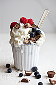A white cup filled with vanilla ice cream, topped with chocolate peanut butter candies, raspberries and blueberries