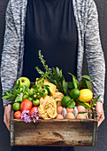 A woman holding a wooden crate of fruit, vegetables and other foodstuffs