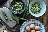 From above of rope fresh green peas in bowl and eggs on wooden table while cooking