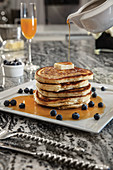 Stack of blueberry buttermilk pancakes with butter adn syrup being poured, paired with mimosa