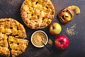 Top view of delicious homemade apple pie with cut slice, fresh apples, peel, cane sugar on brown rustic stone background