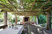 Table and wooden benches below climber-covered pergola with seating area in front of fireplace in background