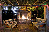 Round table and wooden benches below climber-covered pergola in front of open fire in fireplace