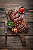 Grilled or fried and sliced marbled meat steak with fork, tomatoes as a side dish and different sauces on wooden cutting board, top view, close-up