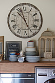 Stacked crockery and bird cage on kitchen counter below large clock on wall