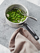 Peas in a saucepan on a grey work surface with a spoon and a cloth