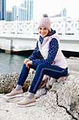 A young woman wearing trousers, a hoodie, a gilet and a bobble hat