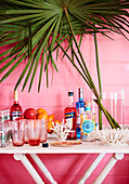 Home bar on white table in front of pink wall decorated with coral and palm fronds