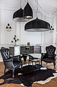 Two black, Baroque armchairs on cowhide rug below large ceiling lamps