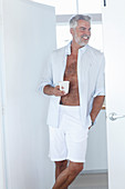 A grey-haired man wearing an open white shirt and white trousers holding a cup of coffee