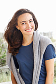 A young brunette woman wearing a short-sleeved blue t-shirt with a grey jumper around her shoulders