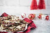 Dunkle und weiße Bruchschokolade mit Cranberries und Pistazien auf Teller, im Hitnergrund Weihnachtsdekoratio