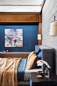 Double bed in front of wooden wall in concrete look in the bedroom, artwork on a blue wall in the background