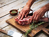 Hands curing a raw fresh angus meat tomahawk on a wooden cutting board with rosemary, salt and butcher's knife