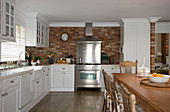 Küche mit weißen Schrankfronten, Edelstahl-Herd vor Backsteinwand und Esstisch aus Massivholz mit Stühlen