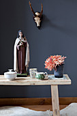 Holzbank mit Blume und Madonna-Figur vor dunkler Wand mit Geweih