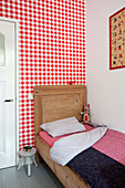 Wooden bed with tall headboard against wall covered in red-and-white gingham wallpaper