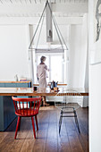 Long dining table with various chairs in kitchen-dining room with woman at window in background