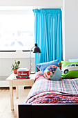Bed with colourful bed linen, bedside table and turquoise curtain in boy's bedroom