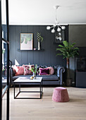 Dunkles Sofa mit Rosa Kissen vor dunkler Wand, Sitzpouf und Couchtisch im Vordergrund