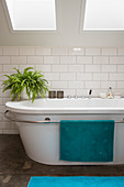 Bathtub with towel rail and indoor fern under skylight in bathroom with white underground tiles