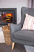 Grey armchair and basket of wood in front of fire in glass-fronted fireplace