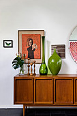 Green glass vases, candlesticks and plants on retro sideboard below artworks on wall