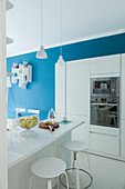 White kitchen cabinets with fitted appliances, counter with bar stools and blue wall