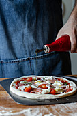 Unrecognizable man in apron spilling olive oil of top of pizza while standing behind table and cooking in kitchen