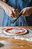 Unrecognizable guy in apron grinding fresh cheese on dough with tomato sauce while preparing pizza on table
