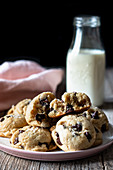 Plate of tasty vegan cookies with chocolate chips placed on lumber table near blurred milk and cloth against black background