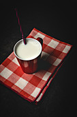Cup of white milk with bright striped straw on table over black background and checkered towel